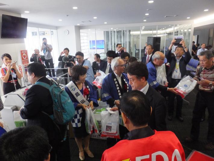 空港にて特産品が入った袋を配る市長たちと、それを撮影する大勢の人たち