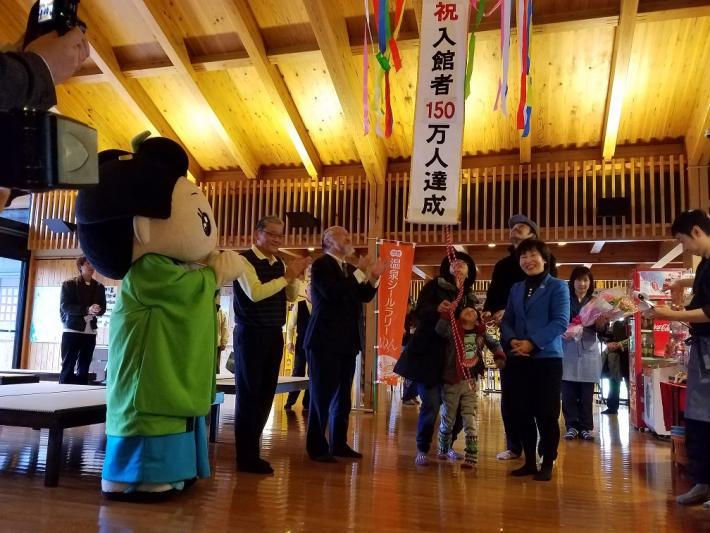 伊太和里の湯にて、女性とこどもが祝入館者150万人達成と書かれたくす玉を割りながら、その周りで市長やゆるキャラ達が祝っている