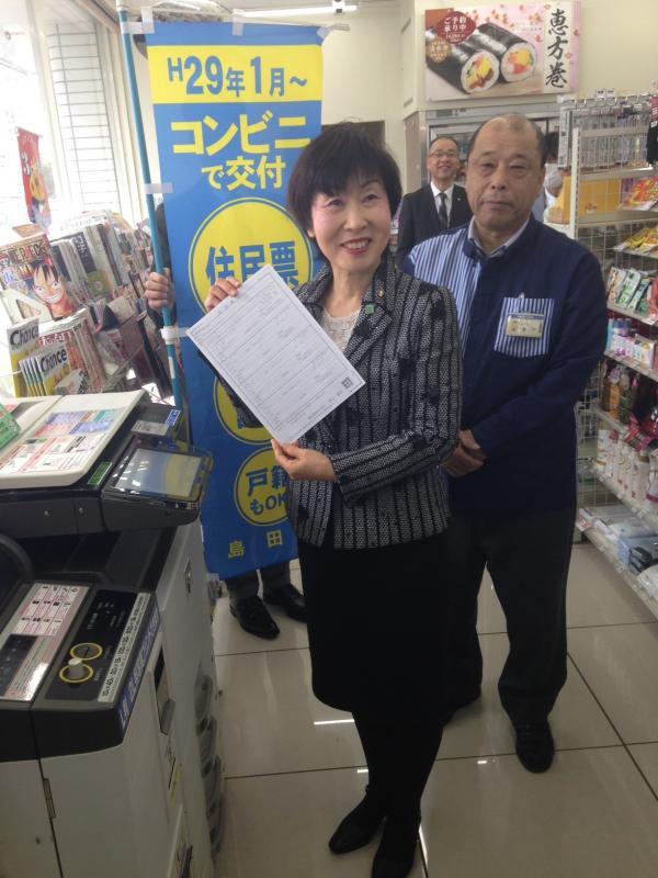 コンビニで住民票等が交付できることが書かれた幟があるコンビニ内のコピー機の前で、書類をこちら見せている市長