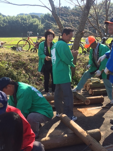 緑の法被を着た市長や男性たちと、丸太で何かを作ろうとしている人たちの和やかな様子