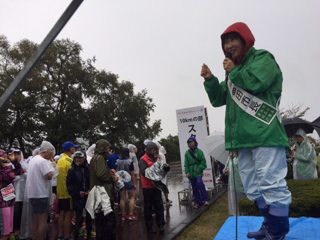 雨の中市長がマイクを持ち、マラソン大会のランナーたちに挨拶している