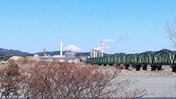 手前に川原と緑の橋、奥に富士山、煙突から煙が出ている写真